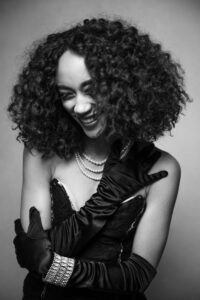 A woman with short curly hair wearing black long black gloves laughing and hugging herselfA woman with short, curly hair is wearing long black gloves, laughing, and hugging herself.
