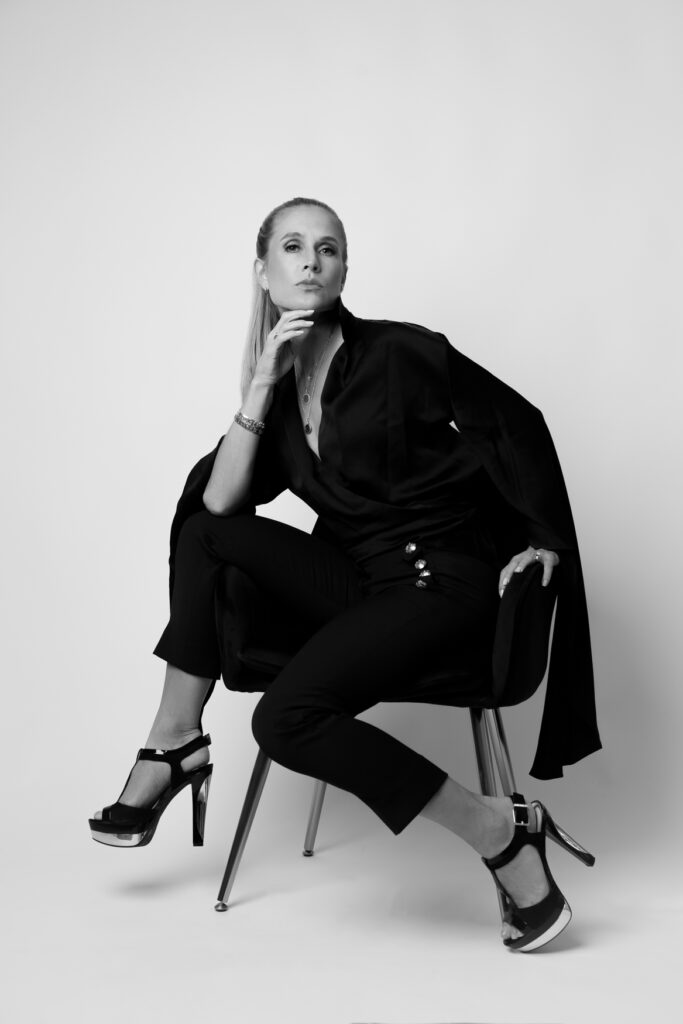 A woman posing confidently to the camera with one hand on her chin, sitting on a chair. 