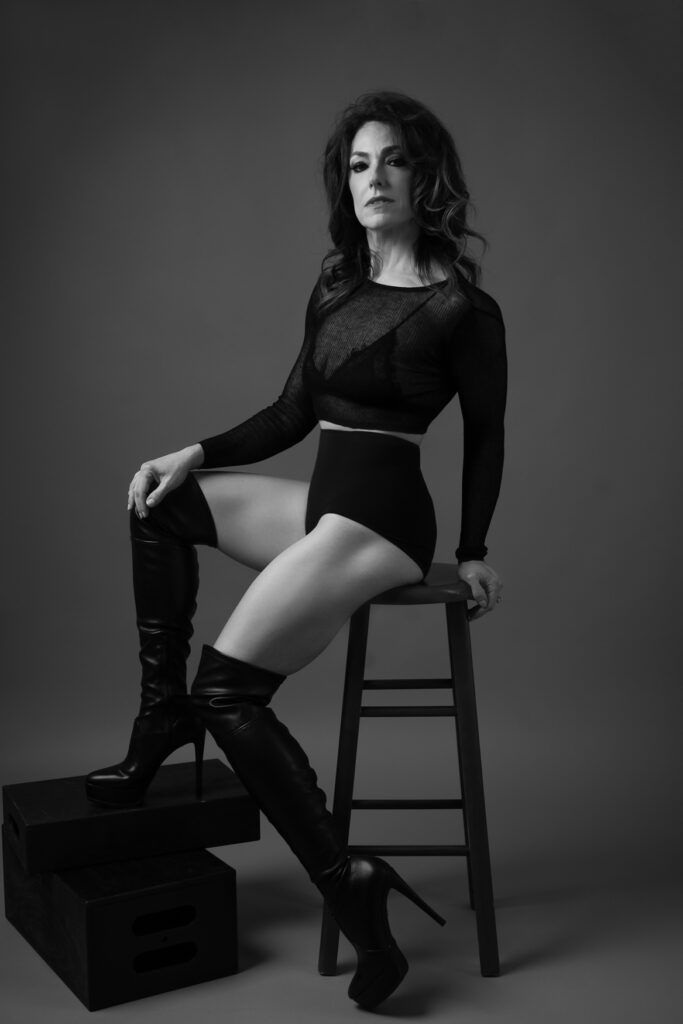 A black and white portrait of a woman sitting on a stool wearing long black boots and black shorts with a black long sleeve top